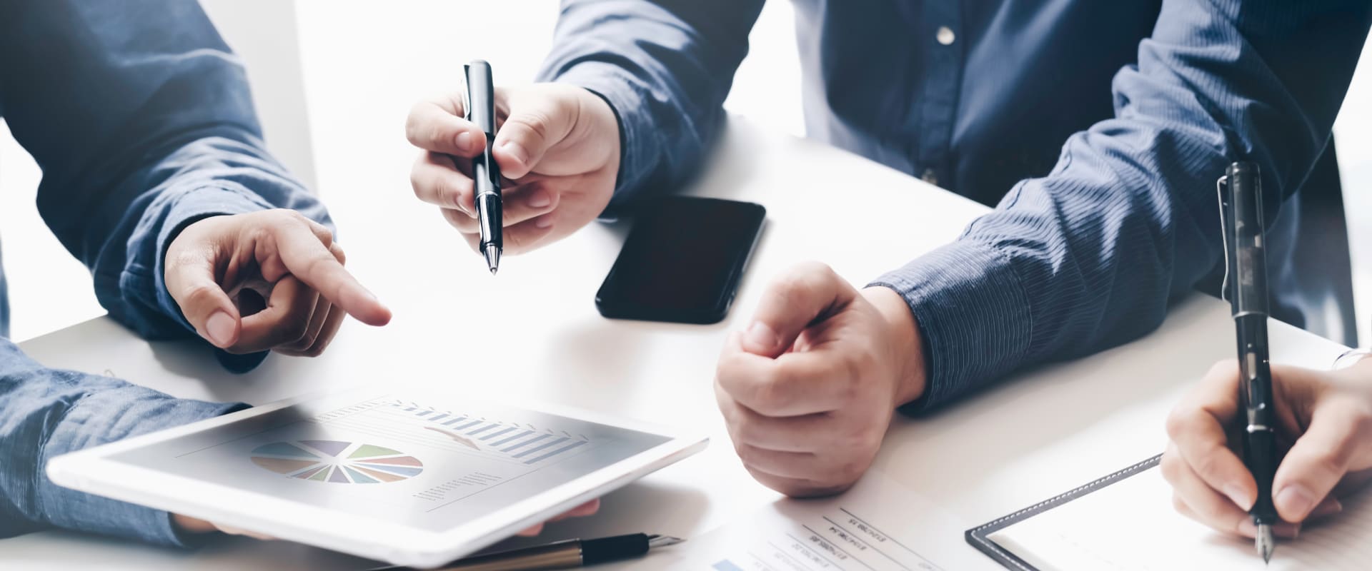 Hände im Business-Meeting zeigen auf Tablet mit Diagrammen, Smartphone am Tisch
